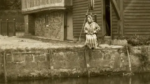 A sepia-tinted photo showing a woman dressed in medieval clothes sitting on a wooden chair which is being lowered over a riverbank to demonstrate how a ducking stool would have worked. An old barn-like building can be seen in the background.