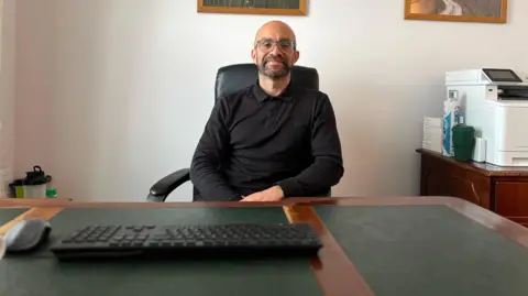 Jason looks at the camera as he he is sat behind a desk. He is wearing a black jumpers with a collar and glasses. Jason has stubbly facial hair and his arms are slightly crossed. There is a keyboard and mouse in front of him and a printer on a cabinet to his right. 