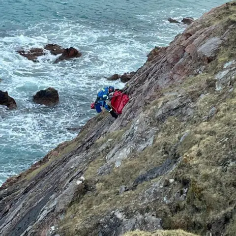 HM Coastguard A man in blue Coastguard colours can be seen down the side of a cliff using a rope, with a red bag that is being used in the rescue of a dog, and the sea can been seen in the background.