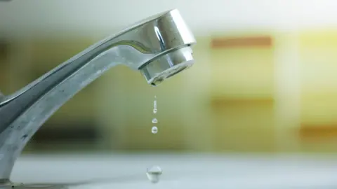 The image shows a tap head with some water drops coming from it. The background is yellow and blurred.
