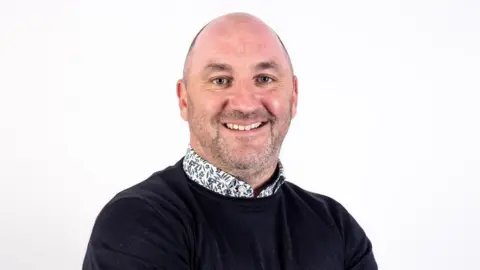 York St John University A man with a bald hear wears a black jumper and floral shirt underneath. He is standing against a white background and smiling at the camera.