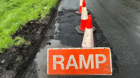 BBC A road with a massive pothole, grass on the left with thick mud and a fluorescent orange sign that says 'RAMP' in capital letters on it in front of fluorescent orange and white cones. 