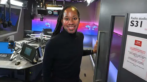 Headshot showing Professor Jason Arday looking into the camera smiling, standing inside the BBC's Today programme studio. He is a black man with long black locs, ear piercings, wearing a black polo neck.