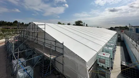 Marquee-style tent covered in white plastic with scaffolding at the nearest end and two-tier water tanks placed along its sides.