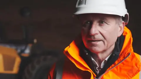 BBC/Becky Holmes James McCoy wearing a white hard hat with orange high visibility jacket