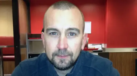 BBC A headshot style picture of David Haines against a red background. He is looking at the camera. He has a shaved head and short beard.