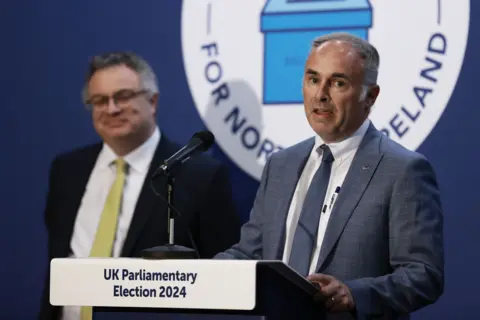 PA Alex Easton in blue suit, giving speech at count centre after 2024 general election. Stephen Farry stands in background in black suit, white shirt and yellow tie