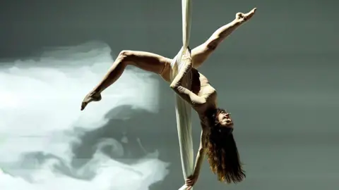 A female performer hangs from a sheet which is wrapped around her body. She is upside down and her legs are open and toes pointed. She has long hair.