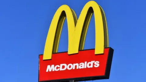 McDonald's sign, featuring large yellow "golden arches" and white logo on red background, set against a bright blue sky.