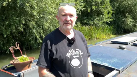 Jeff Slade has short grey hair and is wearing a black t-shirt. He is standing on his canal boat.