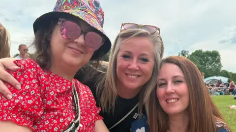 Natasha Rhodes Bryony wearing a red floral dress, a colourful bucket hat and pink-tinted sunglasses on the left of the images, smiling with her sisters Inez - who has blonde hair and a black t-shirt and Natasha, who has red hair. They are all smiling and appear to be at a festival campsite.