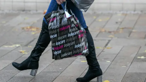 Alamy A person walking on a pavement carrying shopping bags. They are wearing blue jeans and black boots with sparkly heels. One shopping bag is black with the word "bodycare" repeatedly printed in white and pink. Fallen brown leaves are scattered on the ground in the background.