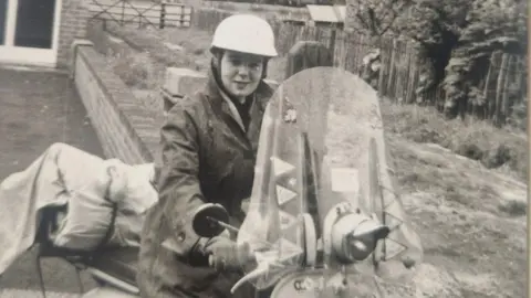 Audrey Morris A black and white image of a woman, Margaret Vickers, sitting on a Vespa scooter wearing a crash helmet and gloves.