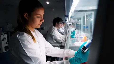 A student working in the new cancer diagnostics laboratory. She is wearing a white lab coat and blue gloves. Behind her are several other students in the same attire.
