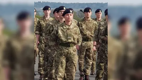 Family photo Gunner Beck wearing her camouflage military uniform and black cap. She is standing among her fellow soldiers with her arms clasped behind her back and a stern facial expression.