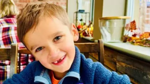 Sheffield Children's NHS Foundation Trust A young boy with short, mousey hair, blue eyes and a gap-toothed smile. He is wearing a blue fleece and an orange T-shirt and sitting on a wooden chair in what appears to be a cafe or restaurant.