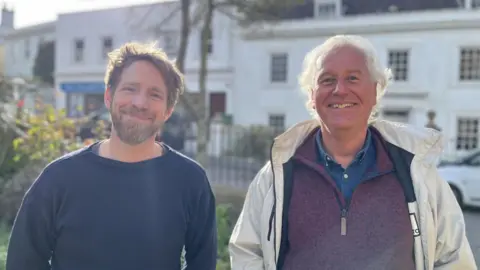 The photo shows Roland and Neil facing the camera in a memorial garden in Alderney. Roland is wearing a navy Guernsey jumper, and has a beard and short brown hair. Neil is wearing a white coat with a mauve quarter-zip jumper underneath, and he has white hair. Both men are smiling.