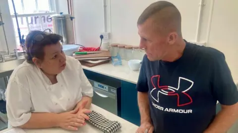 Mousumi Bakshi/BBC Jon being taught how to cook, looking at a woman in a white chef's coat. Jon has a mohawk and a navy "Under Armour" branded T-shirt. The woman he is talking to has short brown hair and glasses resting on top of her head.