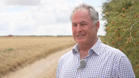 A balding grey haired man smiles while standing in front of a hedgerow laden with red berries. To the left we can see a field of barley recently harvested.