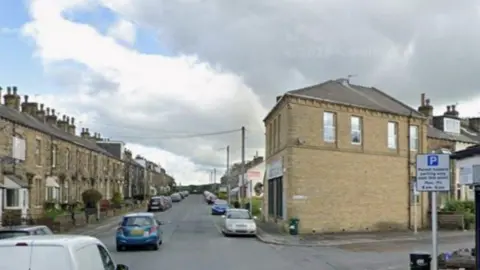 Google Woodhall Terrace in Bradford, with houses and cars and a large yellow brick building on the right.