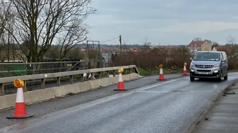 Lewis Adams/BBC Van abu -abu mengemudi di sebelah kerucut di jembatan.