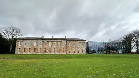 A large two-storey Georgian-style manor house made from yellow stone with a big glass extension on the side.