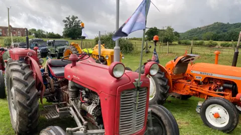 A red tractor has a pole with a flag fixed to the front. The flag has a picture of Harry on the opposite side. There are other vehicles around the tractor in different colours.