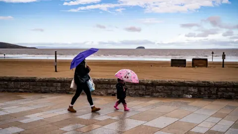 A child with a pink umbrella walks in front of a woman with a blue umbrella on the seafront in Weston-super-mare. The beach and sea are in the background and the sky is cloudy.
