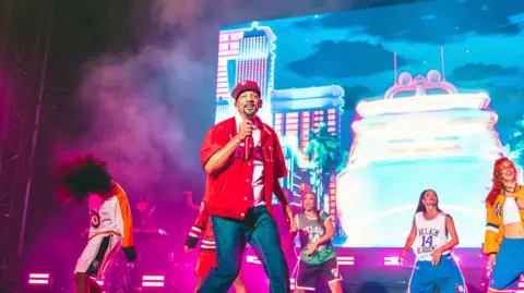 A picture of the actor and rapper Will Smith on the Open Air Theatre stage wearing a red shirt and blue jeans and surrounded by dancers.