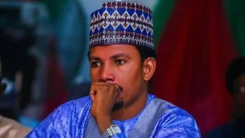 Elisha Abbo Elisha Abbo, sitting with his hand over his mouth, wearing a traditional blue cap and gown