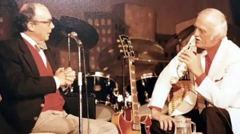 Roger Stennett Eric Morecambe sits on a stage, surrounded by music instruments such as a guitar and a drum kit, as he turns to speak into a microphone on a stand to an older man, who is speaking into a microphone. Morecambe is wearing dark glasses, beige bow tie, red cardigan, brown blazer and cream trousers, and the other man is wearing a white jacket, a red shirt, white tie and dark brown trousers.
