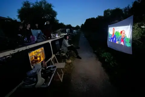 Jan Gazda A sheet set up as a cinema screen along a towpath with people sitting on plastic chairs at the side of a boat watching a movie