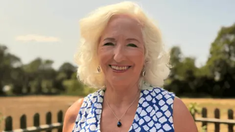 Debbie Taylor wearing a blue-and-white patterned sleeveless top, standing outdoors in front of a fence, field and trees.