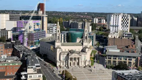 Image shows an ariel shot of Leeds Civic Hall with Millenium Square. 
Behind is a Leeds University glass building with a sky line scene.
To the left of the shot is the Leeds General Infirmary.