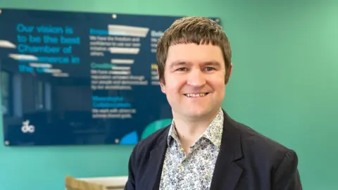 Dan Fell smiles at the camera. He is wearing a dark blazer and a paisley shirt.