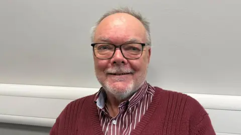 The photo shows a man smiling at the camera. He's wearing black framed glasses, and a maroon jumper with a similar coloured striped shirt underneath