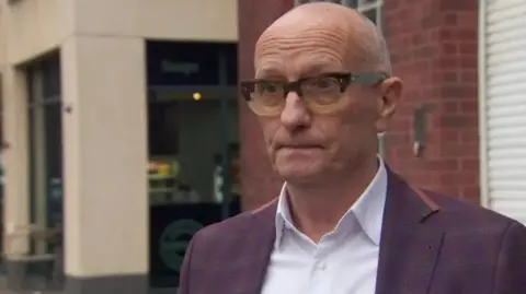 Colin Neill stands in front of  red brick building, he is bald and has on black glasses. He wears a purple tartan suit and white shirt