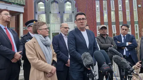 A man in a navy suit with a white T-shirt and dark hair speaks in front of microphones and is flanked by police officers, and politicians and mosque representatives