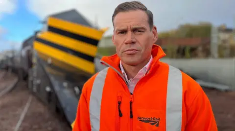 Ross Moran, route director at Network Rail Scotland, stands in front of a snow plough train. He has a serious expression on his face and is wearing an orange hi-viz jacket