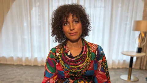 A person wearing a brightly patterned, multicoloured outfit sits indoors against a backdrop of soft curtains and neutral décor. The vivid clothing stands out against the muted room, creating a striking portrait with warm, natural lighting.