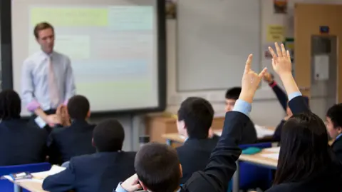 Getty Images Children raise their hands in a classroom as a teacher stands by a white board