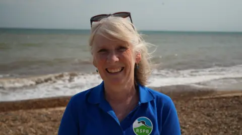 Shaun Whitmore/BBC A woman smiles at the camera in a head and shoulders image. She is wearing a blue polo shirt with a round RSPB badge to the left of her chest, featuring a black and white avocet. The woman has grey hair and sunglasses perched on top of her head. 