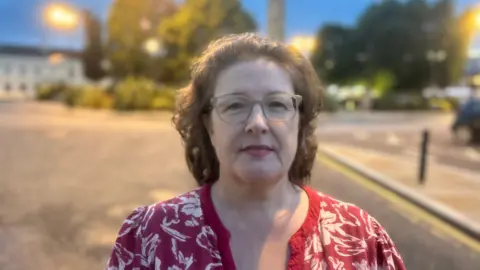 Kellie Armstrong, a woman with shoulder-length brown hair, looks into the camera on a street in Comber. She is wearing grey-rimmed glasses and a red and white top. In the background there are buildings, trees, parked cars and street lights.