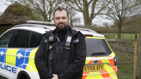 Jamie Niblock/BBC A police officer in a field and standing in front of his police car. He is wearing a his black police uniform, which includes a radio and a body camera. There are trees and a fence behind him.