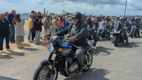 A crowd of people are gathered by a beach to watch a number of motorbike riders drive past.In the centre of the image is a man on a dark motorbike. He is dressed in a suit and has a long moustache. 