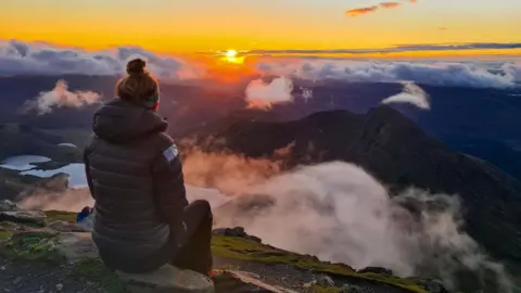 Gemma McInnes A woman is sat on a stone looking at a vast mountainous landscape at sunrise.