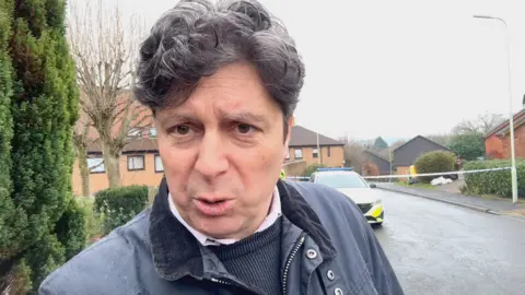 BBC reporter Stephen Fairclough at the scene of the shooting. He is standing on the pavement and is wearing a dark blue coat and navy jumper with a pale pink shirt collar visible. A police car and cordon can be seen behind him on the residential street