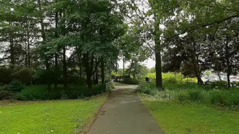 Google Maps A paved footpath runs through a green park with grass and trees on both sides, leading towards a small bridge, with parked cars visible in the distance.
