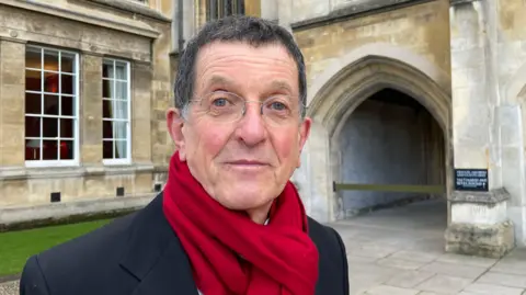 A man wearing a red scarf and glasses is standing in front of a stone building with an archway. There is a large panelled window and a small area of grass behind him.
