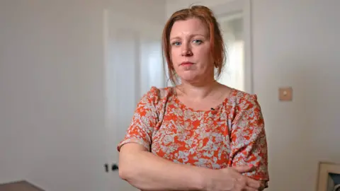 Profile picture of Tassie Weaver. She has straight red hair which is tied back with some tendrils around her face. She is wearing a short-sleeved orange top, patterned with grey flowers and has her arms folded. She is pictured indoors, with a white open door behind her. 